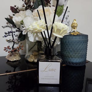 May include: A square dark glass reed diffuser bottle with a white label featuring the gold text Lure, Reed Diffuser, Home Fragrance Redefined, 100 ml. The bottle contains black reeds topped with two cream-coloured fabric flower attachments.