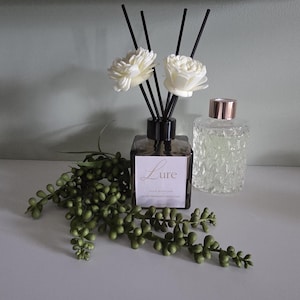 May include: A square dark amber glass reed diffuser bottle with a white label reading Lure, featuring black reeds topped with two white sola wood flowers. Beside it sits a clear textured glass bottle with a metallic rose gold cap.