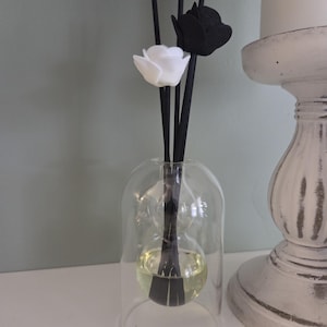 May include: A clear glass reed diffuser with a rounded base containing a pale yellow liquid. Several thin black reeds extend from the top, with one white flower and one black flower attached to the ends of two of the reeds, creating a floral display.