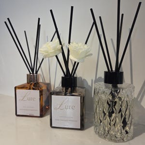 May include: Three reed diffusers with black sticks. Two square glass bottles are amber and dark brown with white labels reading Lure, featuring white flower accents. The third is a clear, textured glass bottle with a black cap and black sticks.