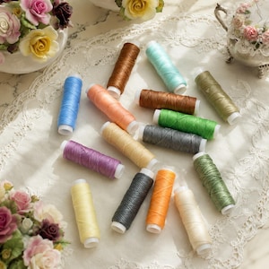 May include: A collection of fifteen cylindrical spools of sewing thread in various colors including blue, brown, peach, light green, dark green, grey, purple, yellow, orange, and cream, each featuring a white plastic core at both ends.