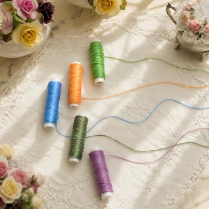 Peut inclure: Cinq bobines de fil cylindriques aux couleurs vives, notamment vert vif, orange, bleu, vert olive et violet. Chaque bobine présente un seul brin de fil assorti s'étendant vers l'extérieur, disposé sur une surface en tissu dentelle blanc.