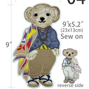 May include: An embroidered sew-on patch featuring a bear wearing a blue jacket, tan pants, and sandals, draped with a colorful patterned blanket. The patch measures 9 inches by 5.2 inches. The image includes a smaller view of the reverse side.