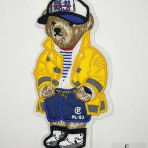 May include: An embroidered sew-on patch featuring a brown teddy bear wearing a yellow raincoat, blue shorts with the text PL-93, a white and blue striped shirt, a white and blue cap with US-93 POLO text, and white sneakers, measuring 7.5 inches by 3.6 inches.