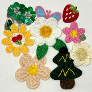 Puede incluir: Una colección de parches de tela variados que incluye una flor de lentejuelas verdes, una margarita amarilla con centro de cereza, una flor blanca con una mariposa azul, una fresa roja, una flor rosa, una flor beige y un pino verde oscuro.