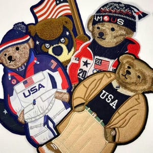 May include: A collection of four embroidered patches featuring brown teddy bears dressed in various outfits. One bear wears a USA jacket, another a hat with the text FAMOUS, a third a superhero mask, and the fourth a USA sweater and tan pants.