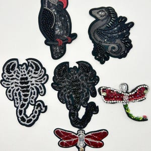 May include: A collection of six embroidered patches featuring a black parrot with a red beak, a dark gray dragon, two scorpions in black and white, and two dragonflies with red wings and clear crystal accents, all detailed with sequins and beads.