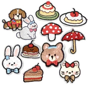 Puede incluir: Una colección de doce parches bordados con varios diseños, incluyendo un perro marrón, un conejo blanco, una seta roja, un paraguas rojo, una cara de oso pardo, una cara de gato blanco y varios postres como pasteles y una porción de tarta.