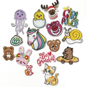 Puede incluir: Una colección de parches bordados termoadhesivos con varios diseños, incluyendo una medusa morada, un pollito amarillo en un huevo, un reno, un unicornio, un caracol, un oso rosa, un conejo con flores, un gato y el texto Girl Power y Be Grateful.