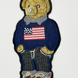 May include: An embroidered iron-on patch featuring a tan teddy bear wearing a navy blue sweater with an American flag design, dark gray pants, and brown shoes. The patch measures 5.1 inches by 2.5 inches and has a black border outline.