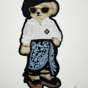 May include: An embroidered patch of a tan bear wearing a black beret, dark sunglasses, a white collared shirt with a small black logo, and a blue patterned wrap skirt over black footwear. The patch measures 7.5 inches by 3.2 inches and is sew on.