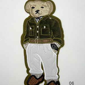 Könnte beinhalten: Ein gestickter Aufnäher in Form eines Teddybären, der ein dunkelolivgrünes Hemd, einen braunen Gürtel und eine hellgraue Hose mit braunen Schuhen trägt. Der Aufnäher misst 22,1 cm mal 9,65 cm und ist mit der Nummer 06 gekennzeichnet.