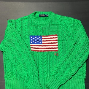 Könnte beinhalten: Ein leuchtend grüner Zopfmuster-Pullover mit Rundhalsausschnitt und einem markanten amerikanischen Flaggendesign auf der Brustmitte. Die Flagge ist in Rot, Weiß und Blau gehalten, mit den Buchstaben RL in rotem Garn unter der Grafik gestickt.