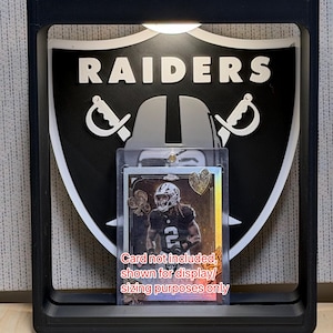 May include: A black rectangular display frame featuring a Raiders shield logo in the center. A clear plastic card holder is positioned in front of the logo, containing a sports trading card with text stating: Card not included, shown for display/sizing purposes only.