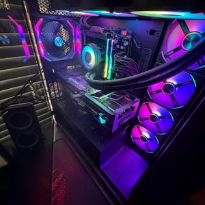 May include: A desktop computer tower with a transparent side panel, featuring internal components illuminated by multi-colored RGB lighting. The system includes a graphics card labeled GeForce RTX, a CPU cooler, and multiple cooling fans.