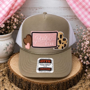 May include: A tan trucker hat with a white mesh back and a foam front. The front features a rectangular pink patch with the text Raisin Hell, flanked by two small brown and pink hearts on the left and a leopard print heart on the right side.