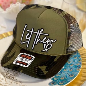May include: An olive green trucker hat with a camouflage pattern on the brim and mesh back. The front panel features white embroidered text that reads Let them with a heart icon. A rectangular sticker on the brim displays the brand name Otto.