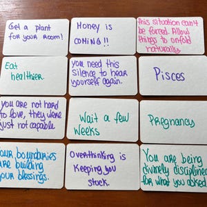 Peut inclure: Un ensemble de douze cartes rectangulaires blanches disposées en grille, chacune présentant des messages manuscrits à l'encre de différentes couleurs. Le texte inclut des phrases comme Get a plant for your room, Money is COMING, Pisces, Pregnancy.