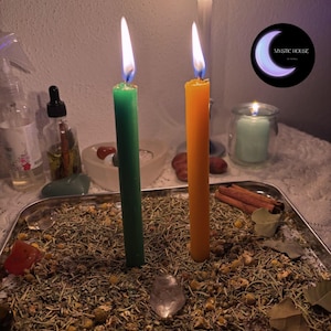 Può includere: Un vassoio metallico rettangolare riempito di erbe e fiori essiccati, con una candela verde e una arancione accese. Un piccolo cristallo trasparente, bastoncini di cannella e foglie secche sono sparsi sul letto di erbe.
