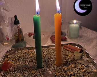 May include: A rectangular metal tray filled with dried herbs and flowers, featuring one green candle and one orange candle standing upright with lit flames. A small clear crystal, cinnamon sticks, and dried leaves are scattered on the herb bed.
