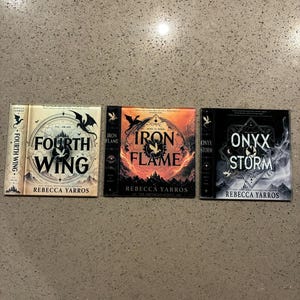Peut inclure: Trois livres reliés de la série The Empyrean par Rebecca Yarros. Le premier, Fourth Wing, a une couverture crème avec des dragons noirs. Le deuxième, Iron Flame, présente un design orange feu et noir. Le troisième, Onyx Storm, est noir.
