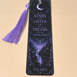 May include: A rectangular bookmark featuring a dark purple and black night scene with a crescent moon, a city landscape, and a bat-like creature. The text reads: TO THE STARS WHO LISTEN AND THE DREAMS THAT ARE ANSWERED VELARIS. It includes a purple tassel.