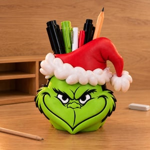 May include: A green pencil holder shaped like a character face with a wide, mischievous grin and narrow eyes. The holder wears a bright red Santa hat with a white textured brim and pom-pom. Several pens and pencils are stored inside the container.