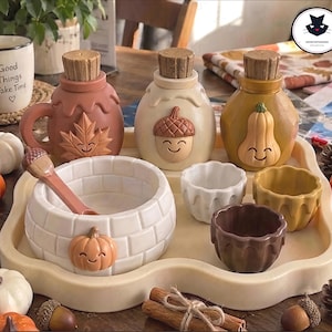 Może przedstawiać: Zestaw ceramiczny składający się z trzech butelek z korkami, dużej miski z łyżką oraz trzech małych filiżanek na tacy. Butelki ozdobione są motywami liścia, żołędzia i dyni. Miska posiada wzór dyni i teksturę cegieł.