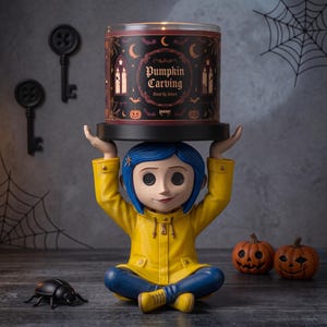 Puede incluir: Un portavelas decorativo con una figura de pelo azul que lleva un impermeable amarillo, sentada con las piernas cruzadas mientras sostiene una gran vela cilíndrica etiquetada Pumpkin Carving y Blood By Values con patrones góticos y calabazas.