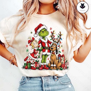 May include: A cream-colored t-shirt featuring a graphic print of a green character in a Santa suit, a blonde girl in a red dress, and a brown dog with antlers. The design includes colorful Christmas lights, wrapped gifts, and green stars.