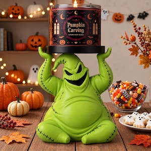 Peut inclure: Une figurine Oogie Boogie vert lime avec des détails cousus noirs tenant un socle à bougie noir. Une bougie cylindrique est posée dessus avec une étiquette noire portant le texte Pumpkin Carving, autumn night candle et des citrouilles.
