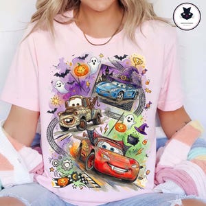 May include: A light pink t-shirt featuring a colorful Halloween-themed graphic of three cartoon cars. The design includes a red race car, a brown tow truck, and a blue car wearing a witch hat, surrounded by ghosts, pumpkins, bats, and candy.