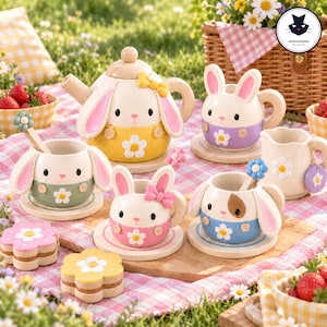 Op de afbeelding: Een keramische theeset bestaande uit een theepot, vier kopjes met schoteltjes, een melkkannetje en twee bloemvormige koekjes. Elk item is versierd met konijnengezichtjes, pastelkleuren en witte madeliefjesmotieven.
