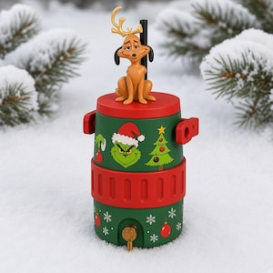 May include: A cylindrical green beverage dispenser with a red lid and base, featuring a brown dog figure with antlers on top. The exterior displays a green character face wearing a Santa hat, a Christmas tree, and red ornaments with white snowflakes.