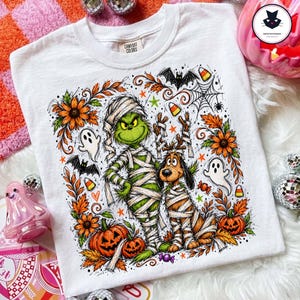 Peut inclure: Un t-shirt blanc avec une impression graphique représentant un personnage vert et un chien marron, tous deux enveloppés de bandages blancs comme des momies. Le motif inclut des citrouilles orange, des tournesols, des fantômes et des chauves-souris.