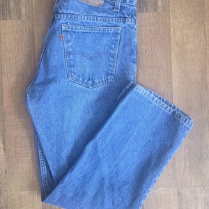 May include: A pair of medium-wash blue denim jeans featuring a classic five-pocket design, a button fly, and a brown leather patch on the back waistband. The back pocket includes a small orange tab with the Levi Strauss and Co brand name.