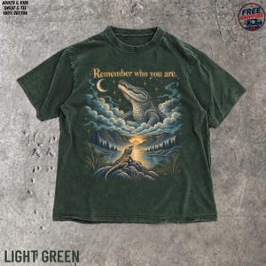 May include: A dark green t-shirt featuring a graphic print of a bearded dragon on a rock looking toward a sunset over water, with a large alligator appearing in the clouds above. The text "Remember who you are" is printed above the alligator.