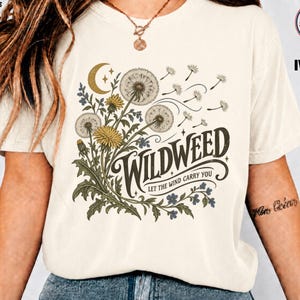 Könnte beinhalten: Ein elfenbeinfarbenes Kurzarm-T-Shirt mit einem Grafikdruck aus gelben und weißen Löwenzahnblüten, einer Mondsichel und kleinen Sternen. Der Text lautet WILDWEED und LET THE WIND CARRY YOU in dunkler Schrift. Das Design zeigt fliegende Samen.