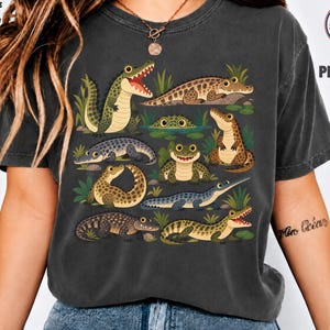 Może przedstawiać: Ciemnoszary t-shirt z nadrukiem graficznym przedstawiającym różne krokodyle i aligatory w stylu kreskówkowym w odcieniach zieleni i brązu, otoczone zieloną trawą i liliami wodnymi. Koszulka zawiera napis Adults & Kids Sweat & Tee 100% Cotton.