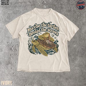 May include: An ivory short-sleeve t-shirt featuring a graphic of two sea turtles swimming in water. The text reads, "maybe the turtles needed the straws." The design includes bubbles and seaweed in shades of blue, green, and brown, with a red and white striped straw.