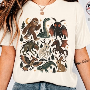 May include: An ivory short-sleeve t-shirt featuring a graphic print of various mythical creatures including a sasquatch, a sea serpent, a moth-like figure, a winged horse, and a jackalope, interspersed with small pine trees and stars.