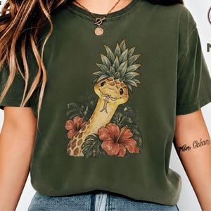 Könnte beinhalten: Ein moosgrünes Kurzarm-T-Shirt mit einem Grafikdruck einer Schlange, die einen Ananas-Aufsatz auf dem Kopf trägt, umgeben von tropischen Blättern und roten Hibiskusblüten. Der Text "ADULTS & KIDS SWEAT & TEE 100% COTTON" und "MOSS" ist sichtbar.