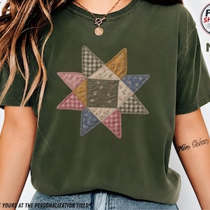 May include: A moss green short-sleeve t-shirt featuring a central graphic of an eight-pointed star composed of various fabric-like patches in muted tones of pink, blue, yellow, and green with checkered and floral patterns printed on the front.