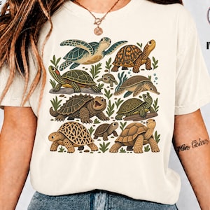 Könnte beinhalten: Ein elfenbeinfarbenes Kurzarm-T-Shirt mit einem Grafikdruck verschiedener Schildkrötenarten in Grün-, Braun- und Beigetönen, durchsetzt mit kleinen grünen Blattillustrationen, die mittig auf der Brust platziert sind.