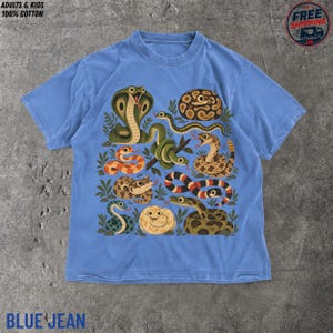 Può includere: Una t-shirt color blu jeans con una stampa grafica di vari serpenti in diversi colori e motivi, tra cui un cobra, un serpente arrotolato e uno a strisce, con il testo ADULTS & KIDS 100% COTTON e BLUE JEAN stampato sull'immagine.