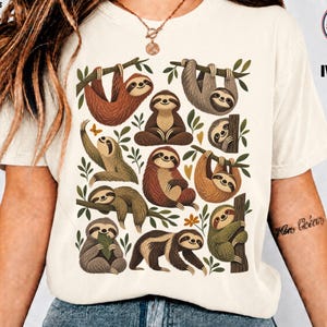 Peut inclure: Un t-shirt ivoire avec une impression graphique représentant plusieurs paresseux dans diverses positions, notamment suspendus à des branches, assis et grimpant. Les paresseux sont représentés dans des tons marron, beige et vert.