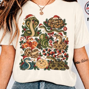 Könnte beinhalten: Ein elfenbeinfarbenes Kurzarm-T-Shirt mit einem Grafikdruck verschiedener Schlangen, die rot-weiße Weihnachtsmützen tragen. Die Schlangen sind von Stechpalmen, Zuckerstangen und Weihnachtsschmuck umgeben. Der Text ADULTS & KIDS 100% COTTON und IVORY ist zu sehen.