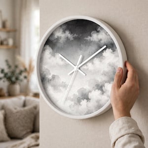 May include: A round wall clock with a white frame and a monochromatic watercolor print of clouds on the face. The clock features three thin white hands positioned against a gradient background of dark gray, light gray, and white cloud formations.