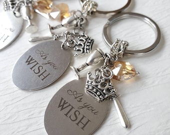 May include: A silver-toned metal keychain featuring an oval pendant engraved with the text As you WISH. The keychain includes a small silver crown charm, a miniature sword charm, a silver goblet charm, and a faceted amber-colored crystal bead.