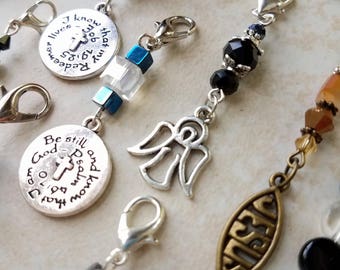 May include: A collection of religious-themed charms and pendants. The charms include crosses, angel wings, and medallions with inspirational text. Various beads in blue, black, and brown are also present. The charms are attached to silver-tone clasps.